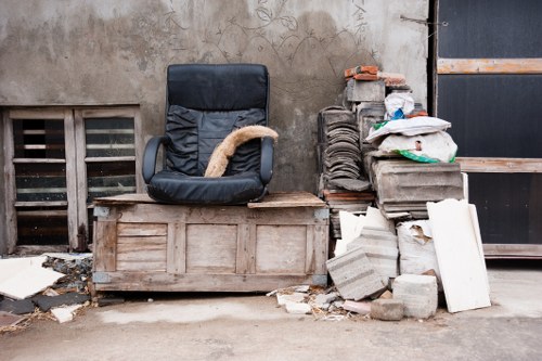 An outdoor scene in an alleyway or industrial area shows a black office or executive chair with a worn, beige fabric armrest resting on a wooden crate or box against a weathered concrete wall. To the right of the chair, there is a pile of construction or renovation debris consisting of grey cement blocks, white plasterboard panels, and broken pieces of concrete, stacked unevenly on the ground. Some loose fragments and smaller debris are scattered on the pavement. The background includes a vent with slats embedded into the wall and a black metal door or shutter on the right side. The environment appears to be part of a waste collection or disposal area, with natural lighting indicating daytime, emphasizing the cluttered state typical of rubbish awaiting removal, possibly by services like Commercial Waste Removal Docklands.