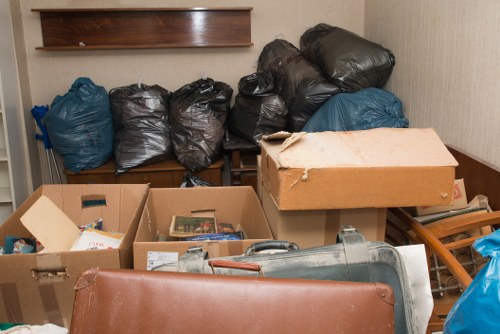 The image shows a cluttered indoor space with multiple black and blue plastic rubbish bags stacked against the back wall, some of which are leaning and appear to be filled with general waste. In the foreground, several open cardboard boxes contain various miscellaneous items, possibly paper or small household objects. A large beige cardboard box, partially torn, sits atop a wooden surface, and nearby, a grey plastic container is visible, possibly a trash or storage bin. To the right, a wooden step stool or small ladder is partially visible, and the environment appears to be a storage or utility room with plain cream-colored walls and minimal natural lighting. This scene illustrates typical waste collection preparation, suggesting services like those offered by Commercial Waste Removal Docklands, in an environment that could be part of a commercial or residential property in the Docklands area, possibly within postcode E14 or a nearby district.