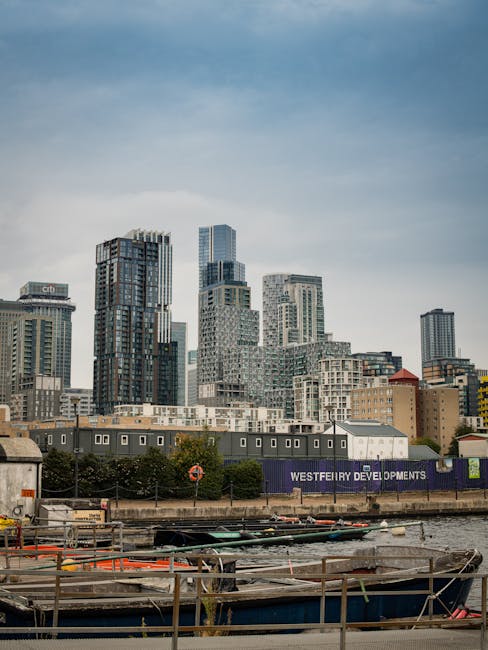 A panoramic view of a modern city skyline featuring numerous tall office buildings and skyscrapers with glass facades reflecting the overcast sky. In the foreground, there is a water body, likely a ri