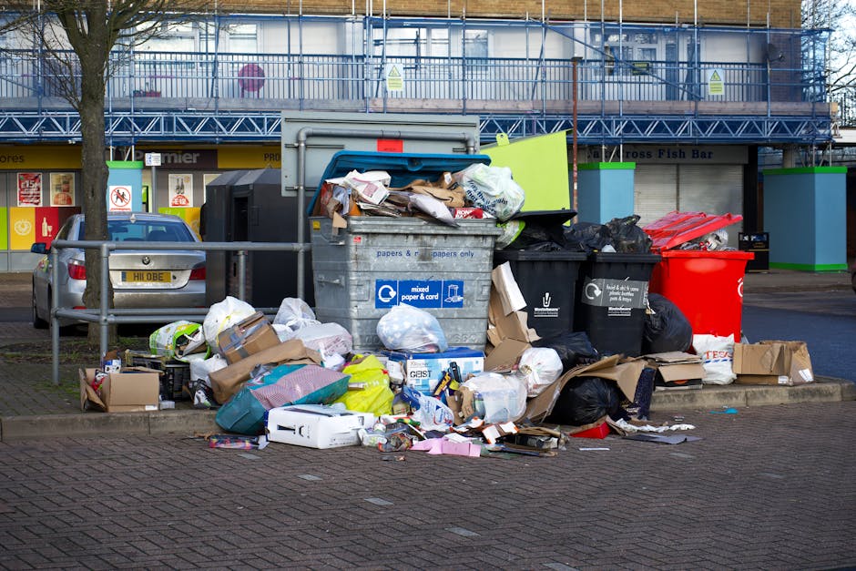 A large, overflowing collection of rubbish and discarded packaging materials piled on a paved sidewalk in front of a commercial or retail area, with various black, red, and brown bin bags along with o