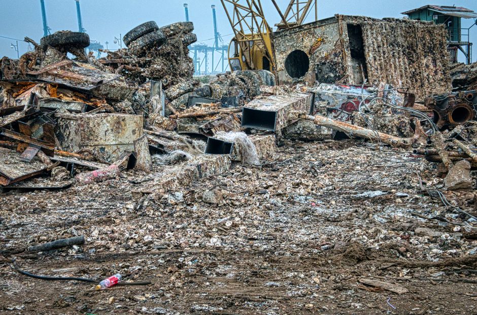 A large, overflowing collection of rubbish and discarded packaging materials piled on a paved sidewalk in front of a commercial or retail area, with various black, red, and brown bin bags along with o